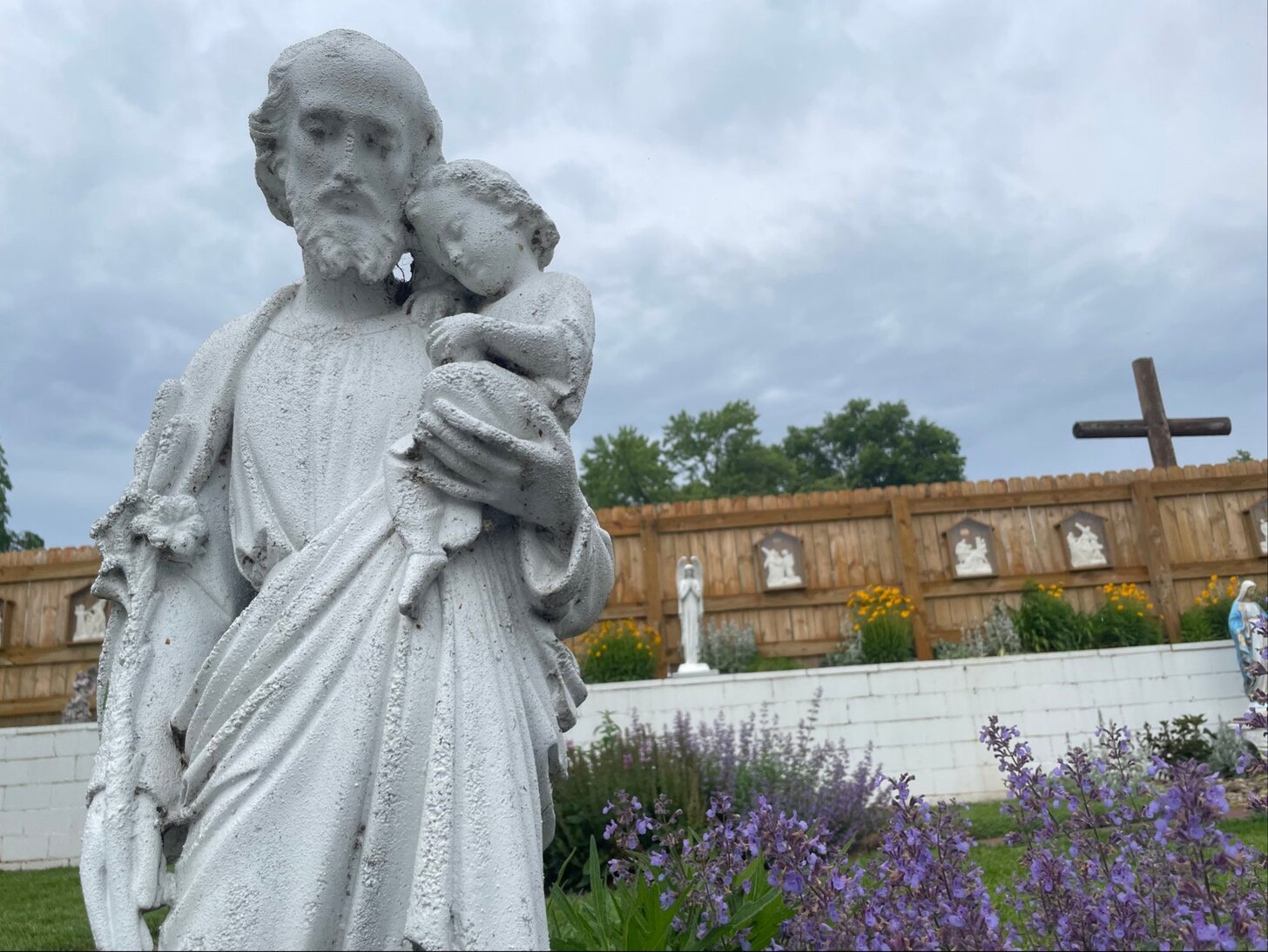 St. Joseph statue in OSMM garden with purple flowers and stations of the cross in the background